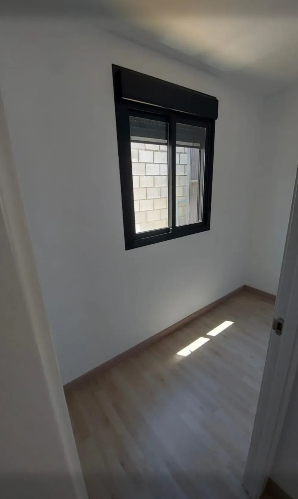 Habitación vacía con una ventana con persianas negras cerradas. La habitación tiene suelo de madera clara y paredes blancas.