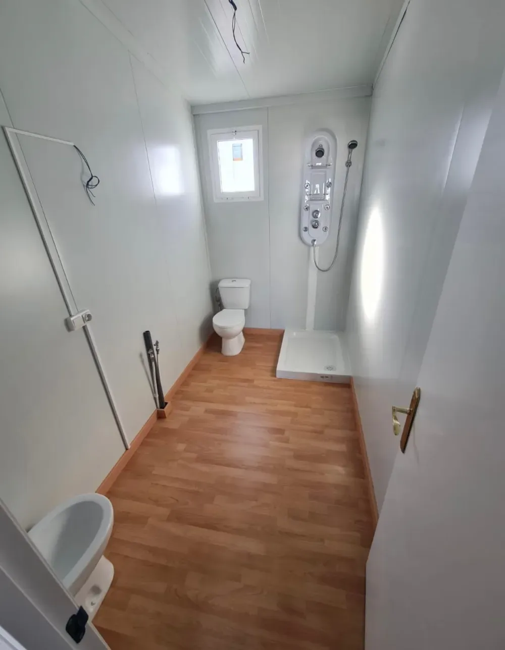 Baño con dos inodoros, ducha y lavabo. Paredes blancas y suelo imitación madera.