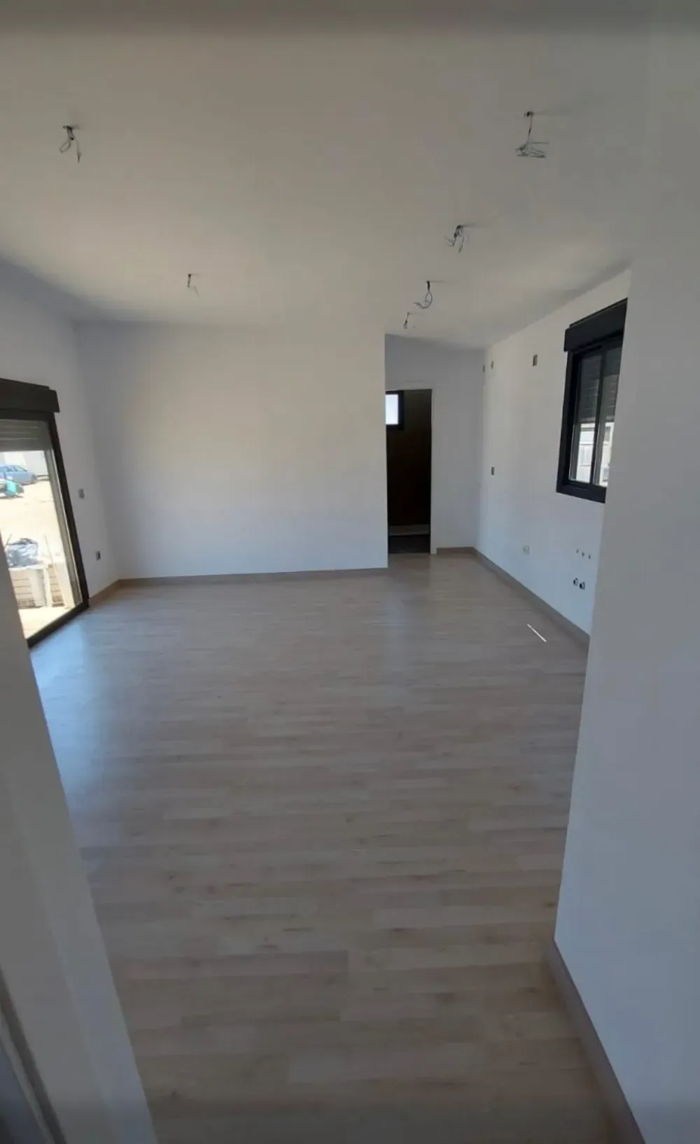 Habitación vacía con pisos de madera clara, paredes blancas, ventanas con marco negro y una puerta.