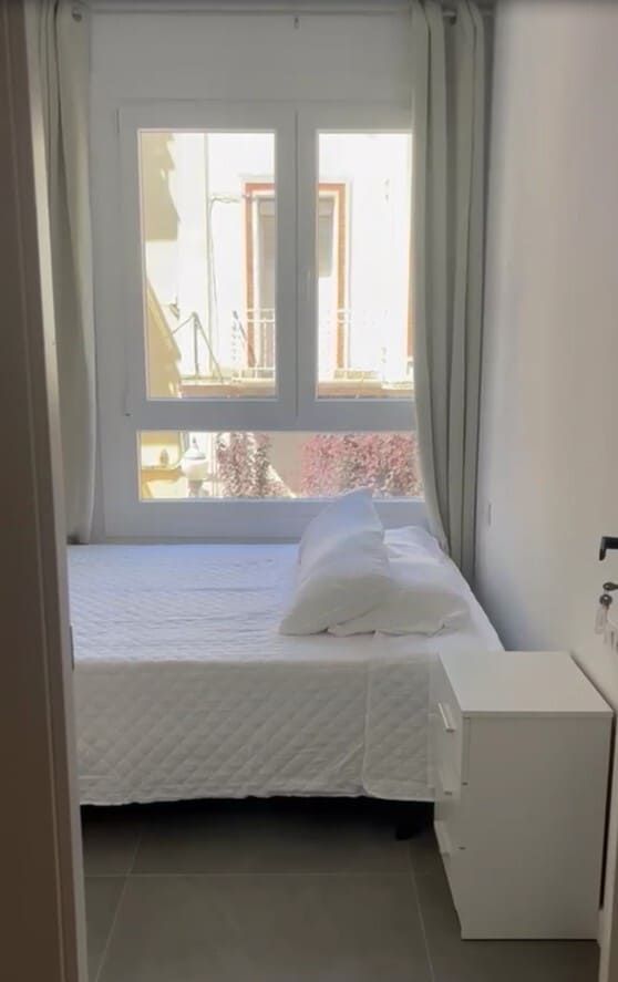 Habitación pequeña con una cama bien hecha, cortinas blancas y una ventana que deja entrar la luz del día.