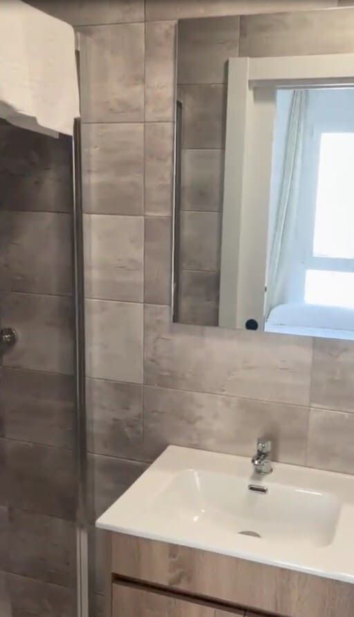 Baño moderno con azulejos grises, lavabo blanco y espejo sobre el tocador.