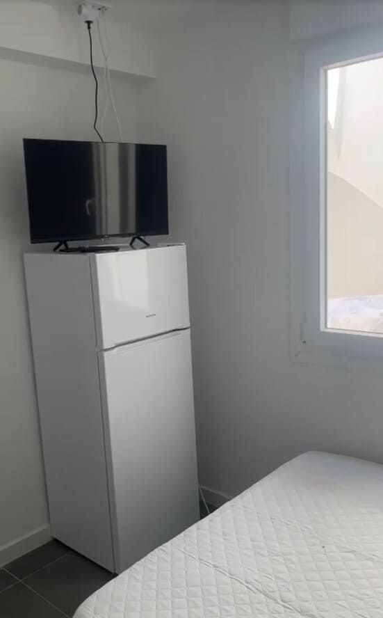 Habitación pequeña de color blanco con nevera y televisor sobre un mueble junto a la cama, cerca de una ventana.