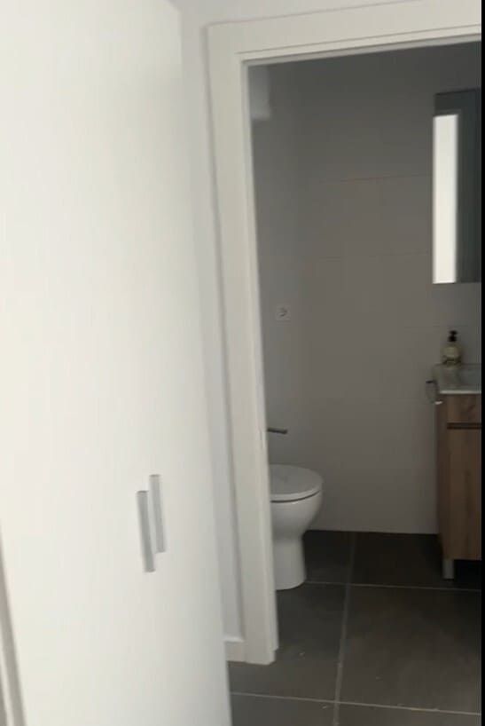 Entrada abierta a un pequeño baño con inodoro, suelo de baldosas oscuras y lavabo de madera.