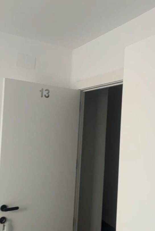 Una puerta blanca abierta marcada con el número “13” en un pasillo sencillo, con una habitación oscura visible al fondo.