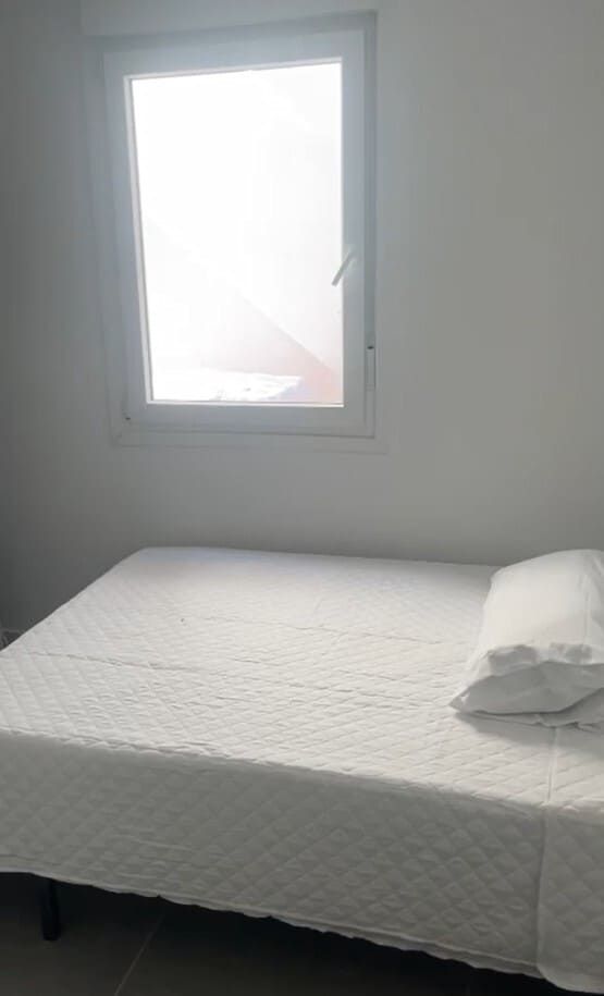 Habitación pequeña con una cama y una almohada blancas debajo de una ventana luminosa.