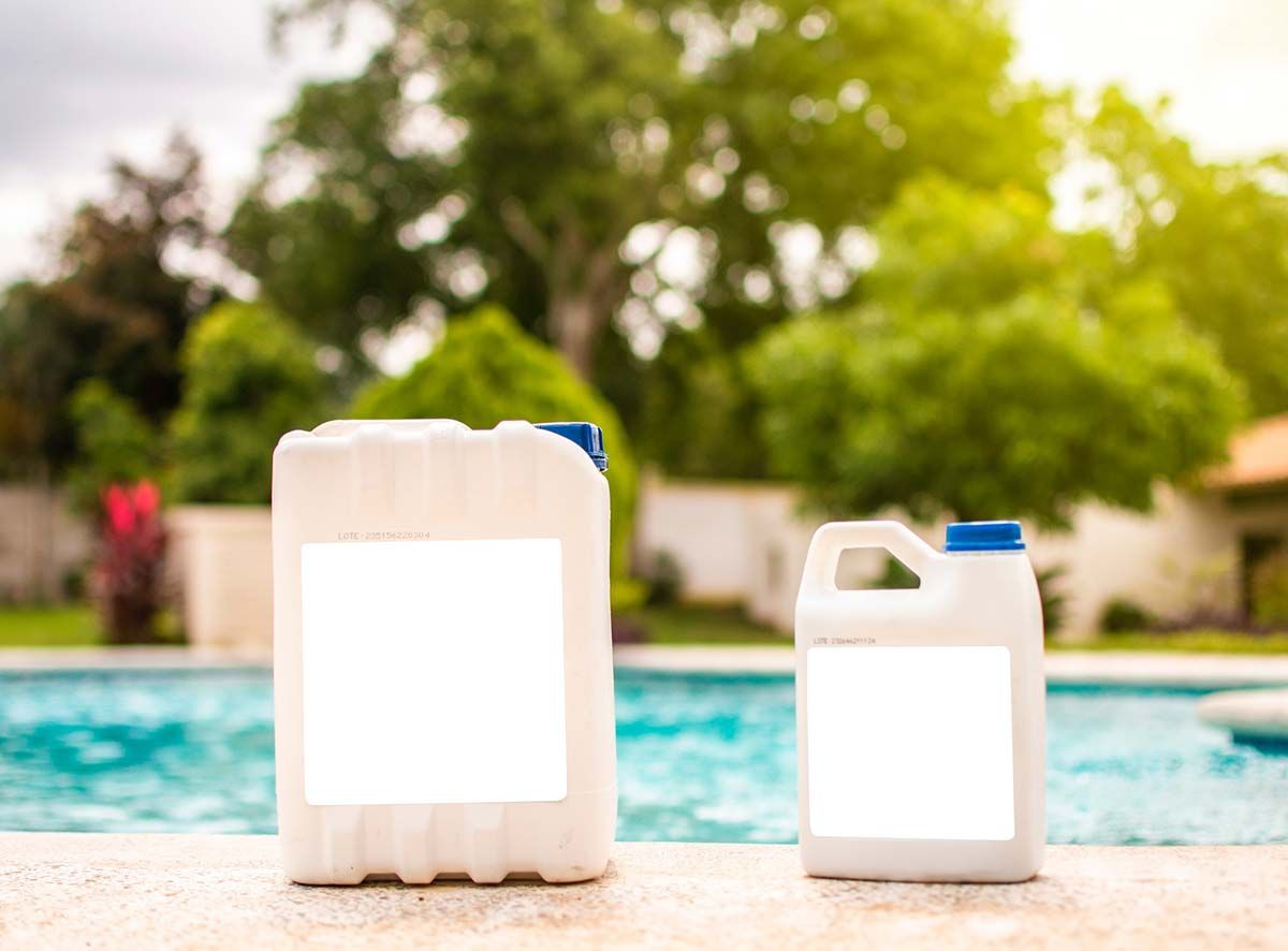 Dos jarras de plástico blancas con etiquetas en blanco se encuentran junto a una piscina, con un fondo verde y borroso de árboles.