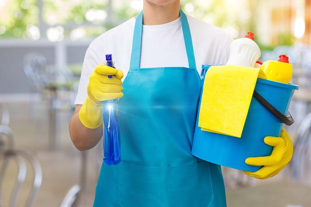 Persona con delantal y guantes, sosteniendo artículos de limpieza: botella de spray, balde con paños y botellas.