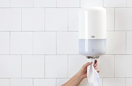 Una persona sostiene un rollo de papel higiénico frente a un dispensador de papel higiénico.