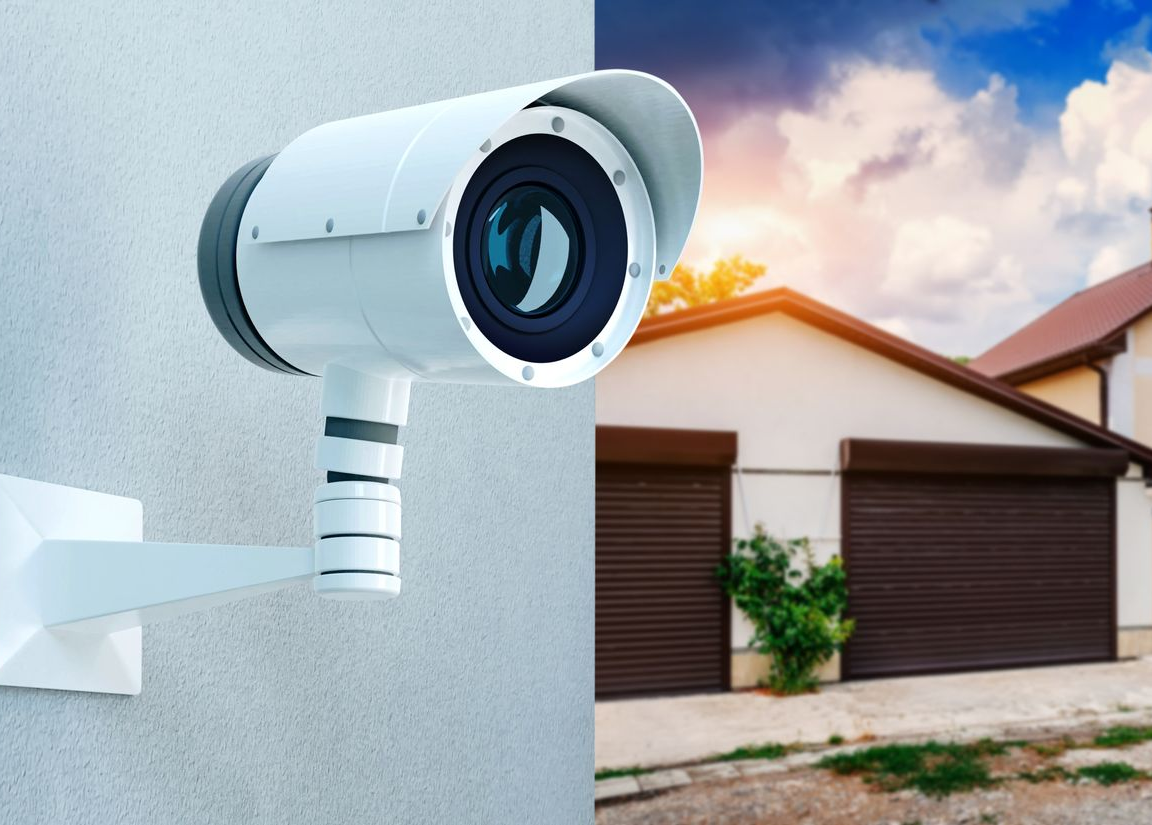 Caméra de sécurité blanche fixée au mur, pointée vers une maison avec des portes de garage.