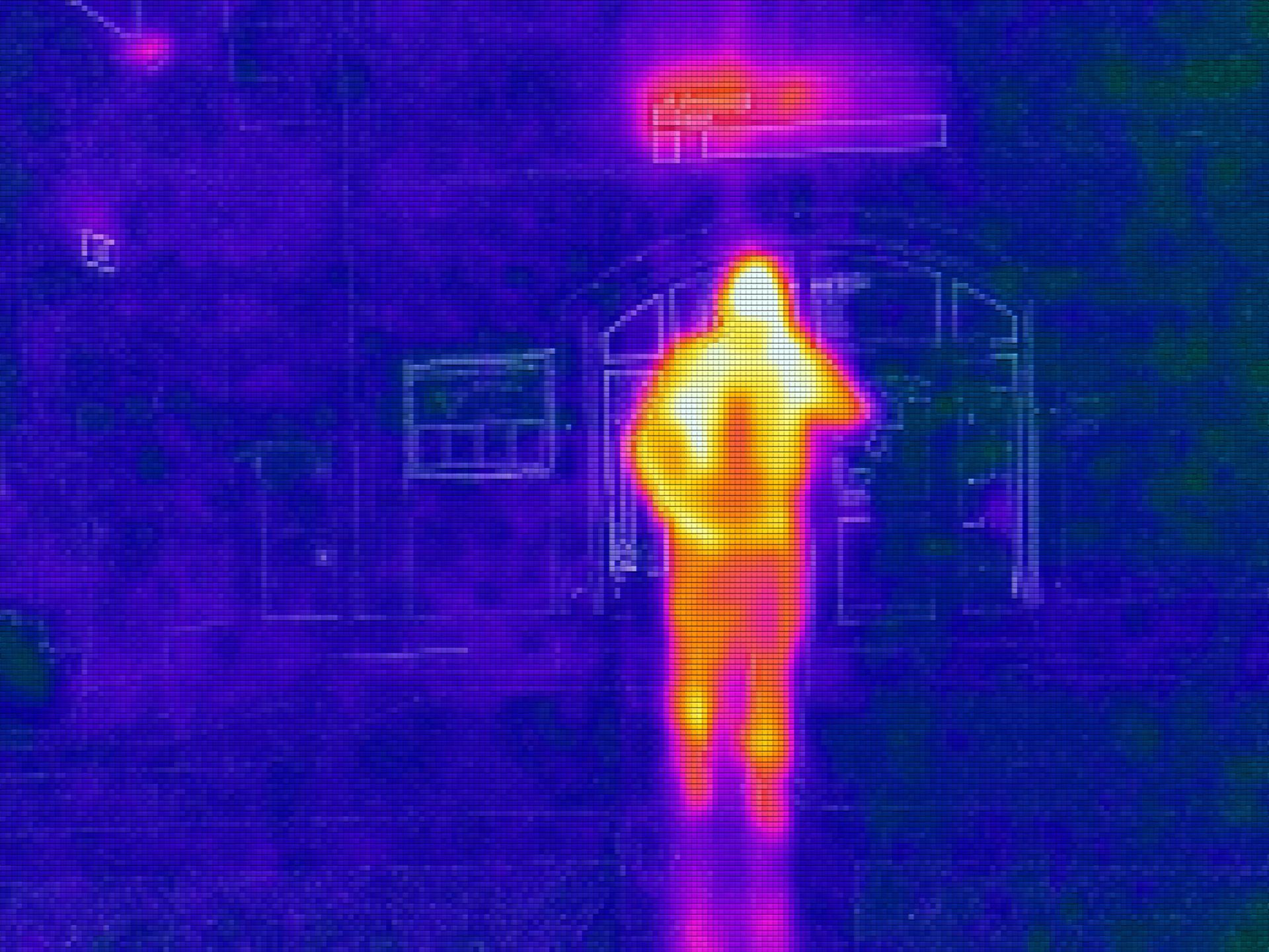 Image thermique d'une personne debout devant un bâtiment, aux couleurs vives jaunes et oranges sur fond violet.