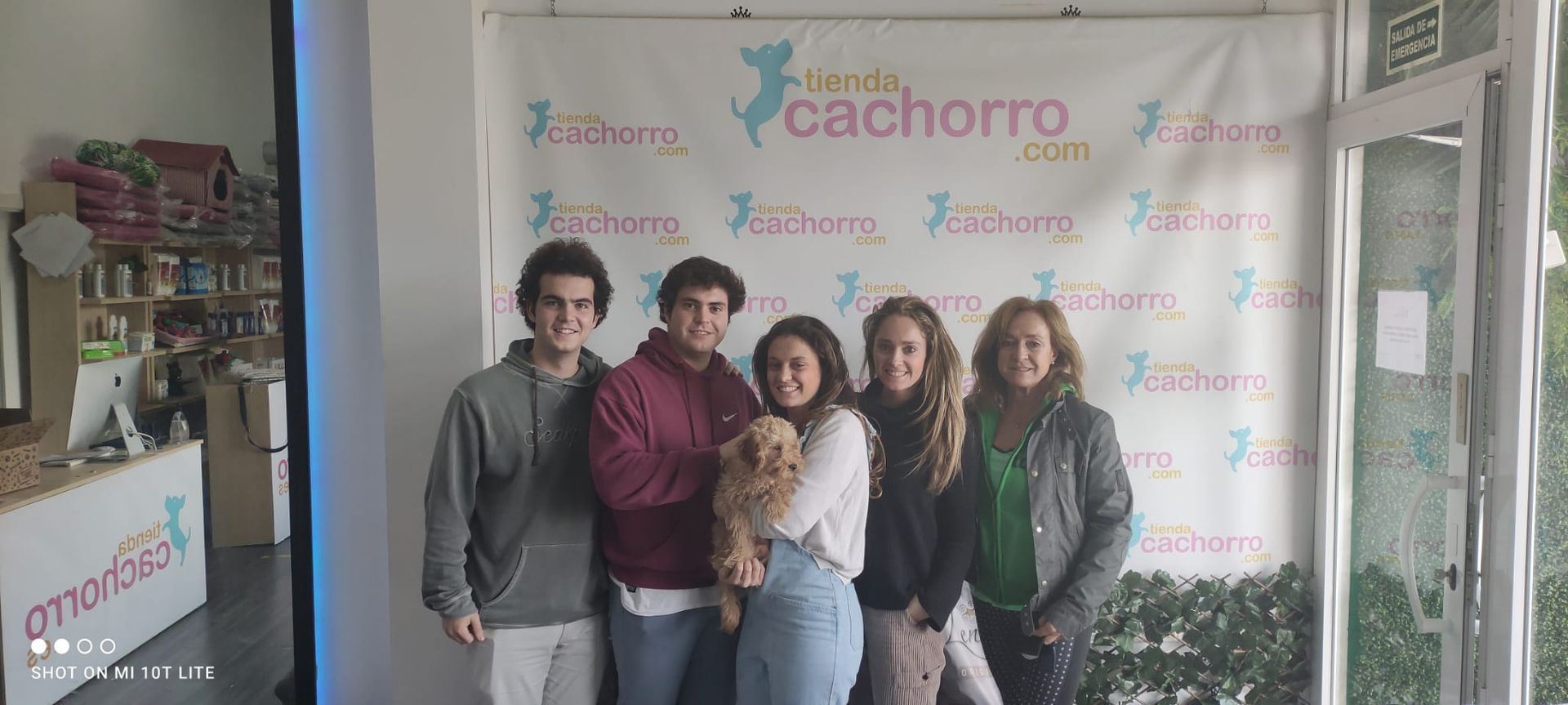 Cinco jóvenes adultos y un perro pequeño posan frente a una pancarta; el escenario parece ser una tienda de mascotas.