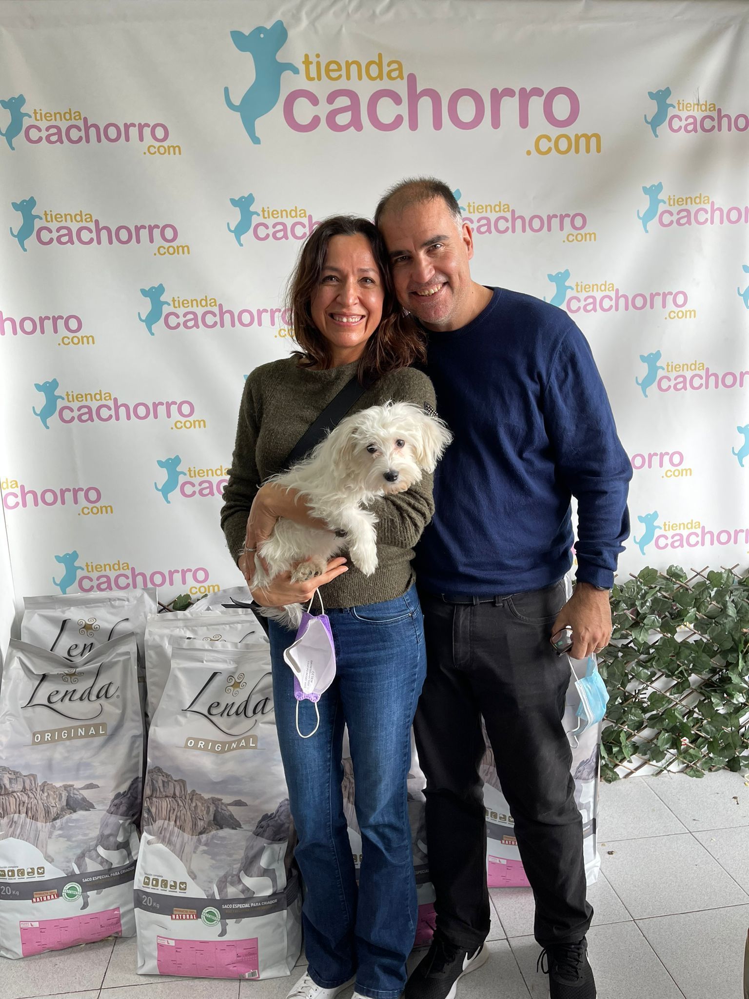 Una pareja sosteniendo un cachorro blanco y esponjoso posando frente a un fondo de "tienda cachorro".