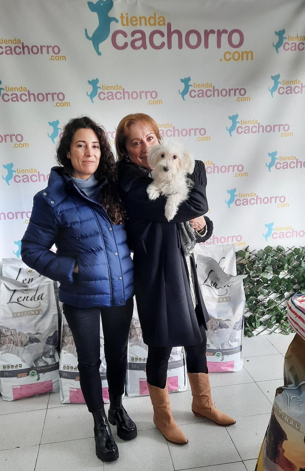 Dos mujeres con un pequeño perro blanco posan frente a una pancarta de "tienda cachorro", rodeadas de bolsas de comida para mascotas.