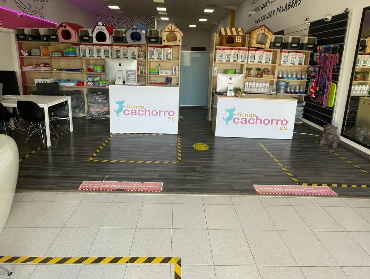 Dentro de una tienda de mascotas: mostradores, estantes con suministros, camas para perros. Marcas de distanciamiento social en el suelo.