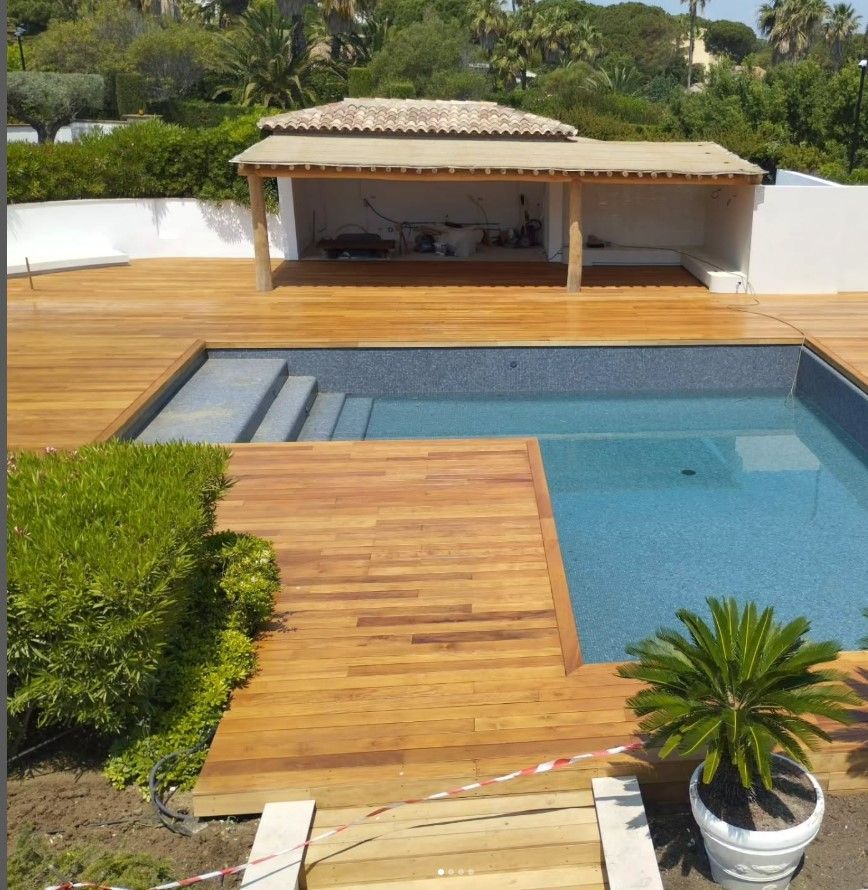 Une terrasse en bois entoure une piscine carrelée de bleu, avec une petite structure couverte et des plantes vertes.