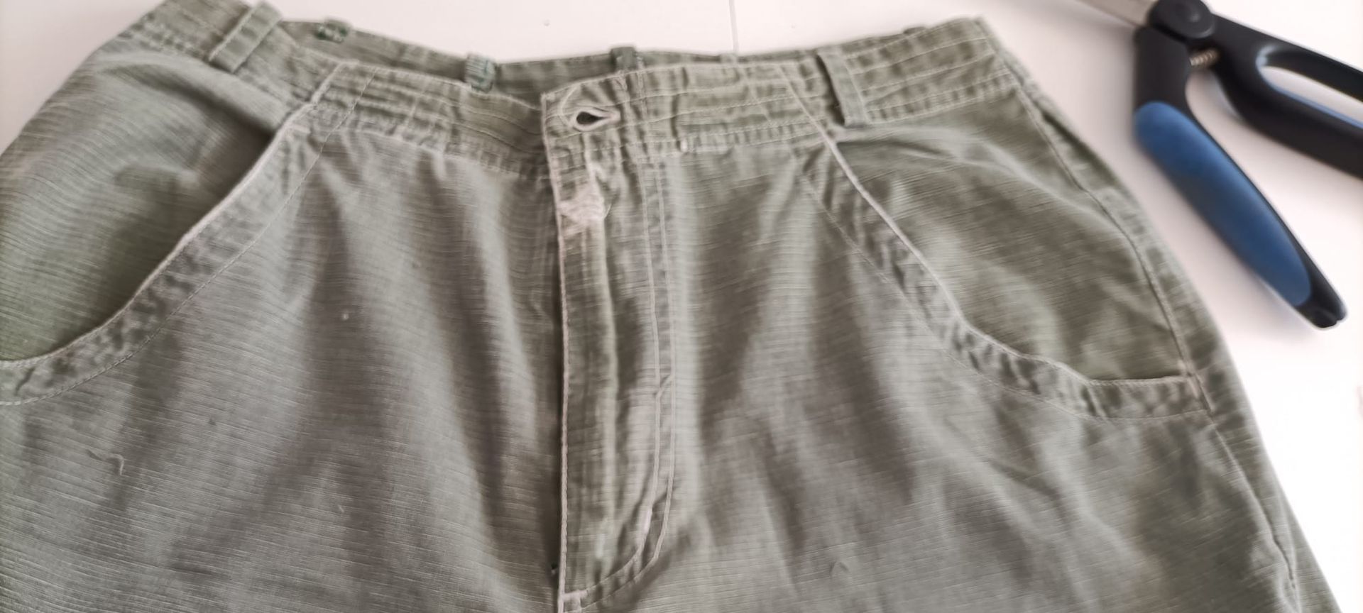 Pantalones cortos de color verde oliva con cierre de botones y bolsillos delanteros, expuestos junto a unas tijeras sobre una superficie blanca.