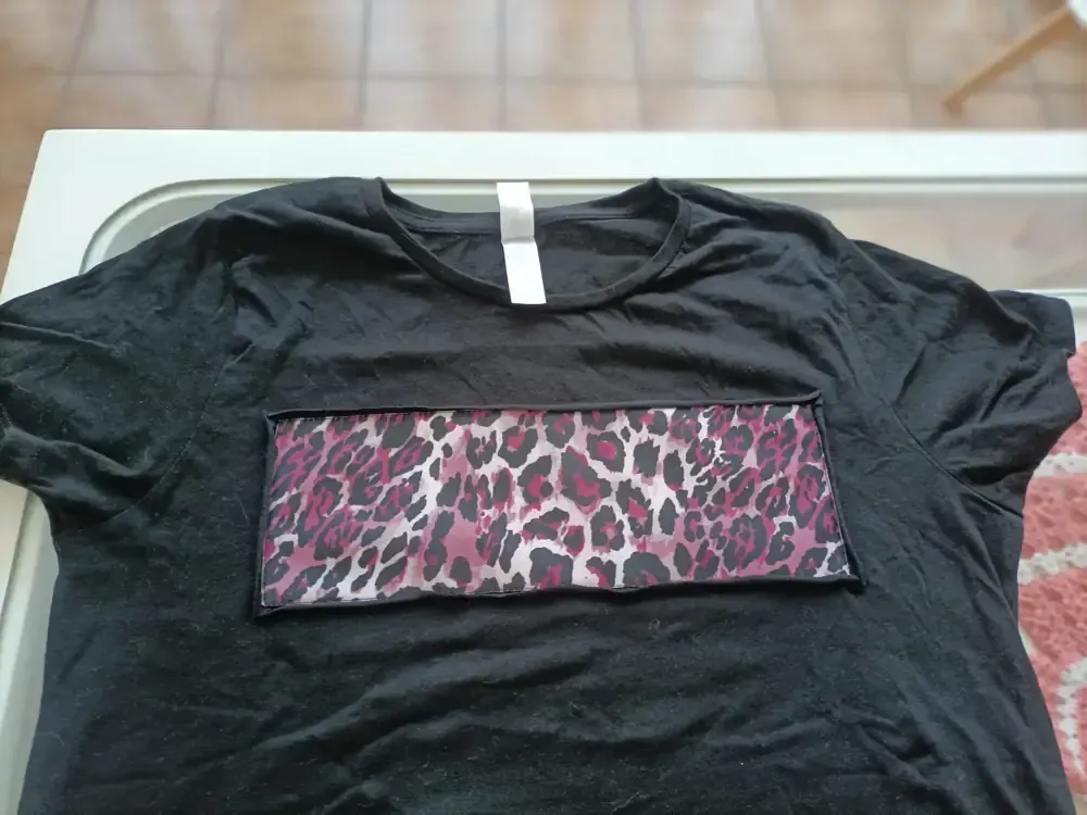 Una camiseta negra con un parche rectangular con un diseño de estampado de leopardo rosa y negro en la parte delantera.