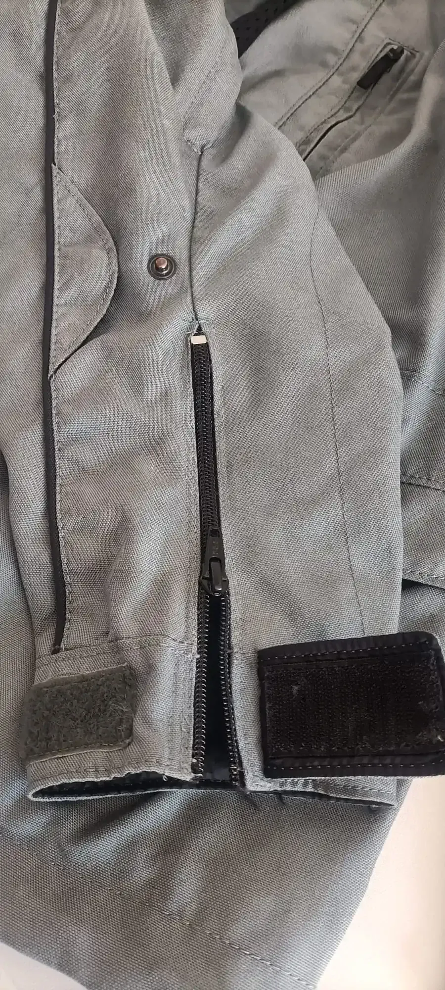 Primer plano de la manga de una chaqueta de moto gris texturizada, con cremallera negra, botón a presión y lengüetas de cierre de velcro.