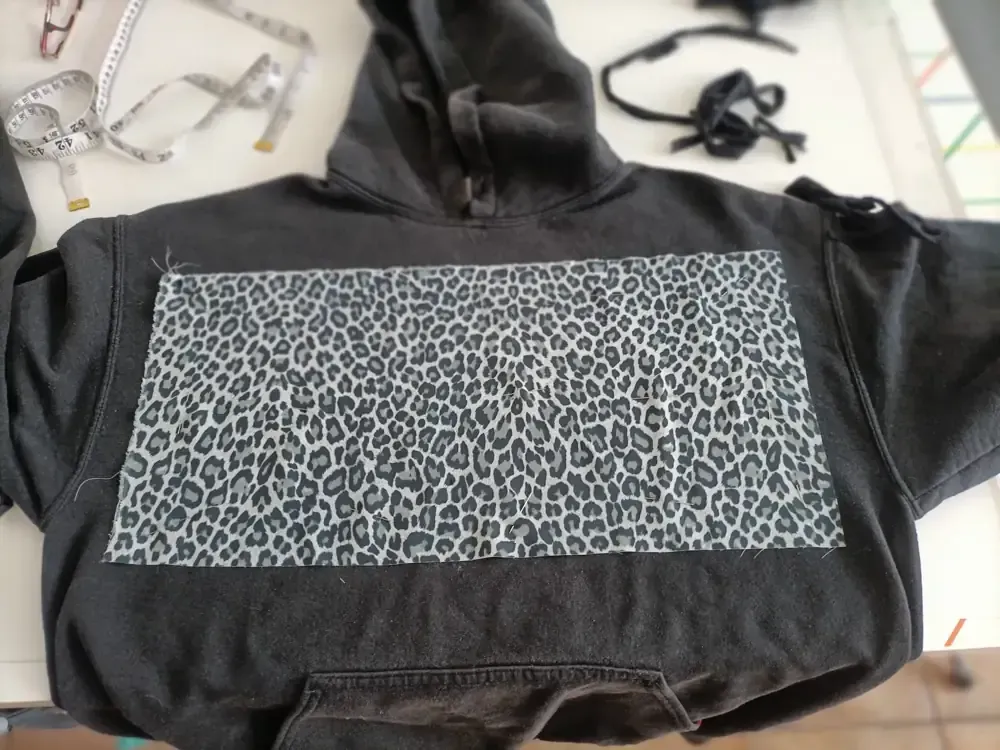 Una sudadera con capucha negra que presenta un parche rectangular de tela gris con estampado de leopardo cosido en el centro del pecho.