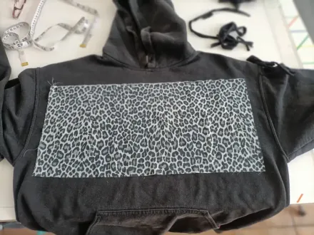 Una sudadera negra con capucha y un gran parche rectangular de tela con estampado de leopardo gris y negro cosido en la parte delantera.