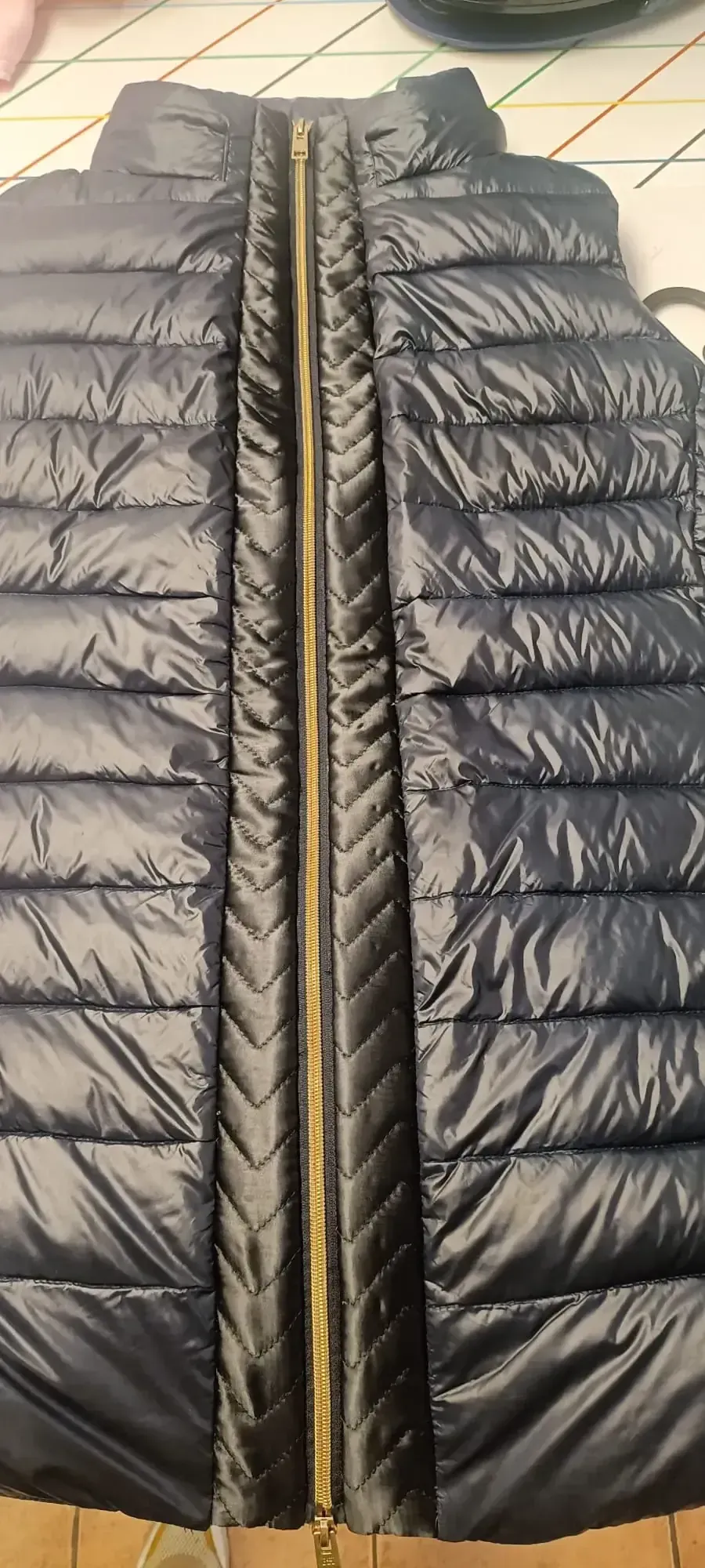 Un chaleco acolchado de plumas azul marino con cremallera central dorada, que se muestra extendido sobre un suelo de baldosas.