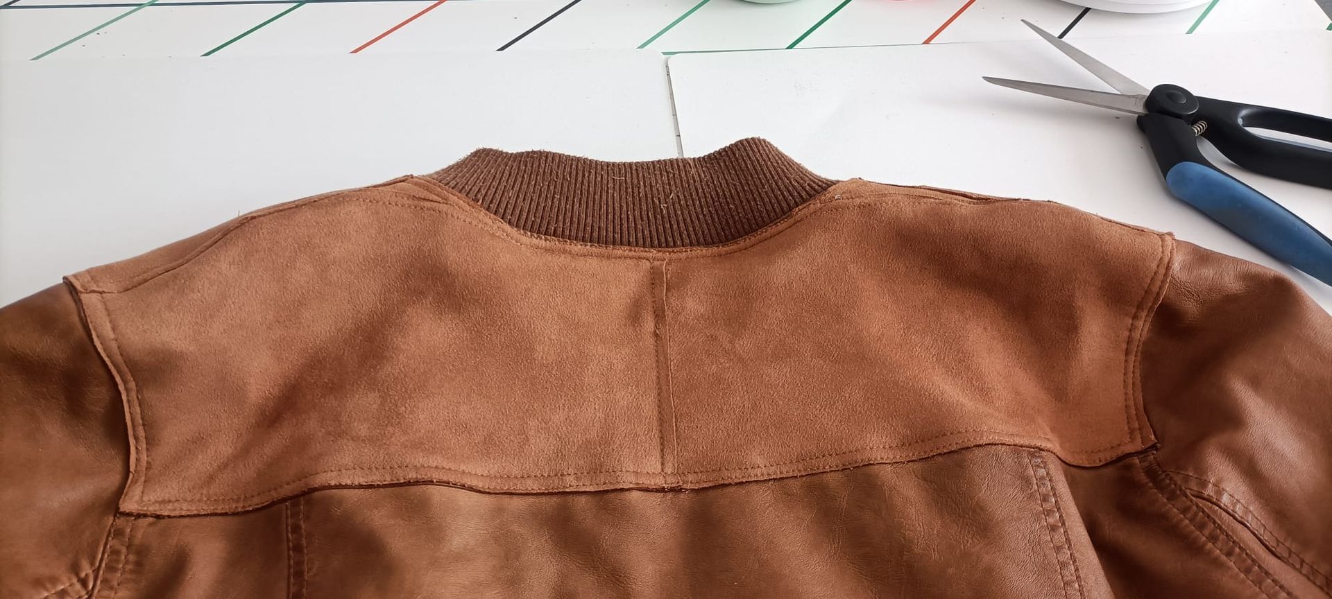El cuello y la zona de los hombros de una chaqueta bomber de ante marrón descansan sobre una mesa blanca junto a unas tijeras.