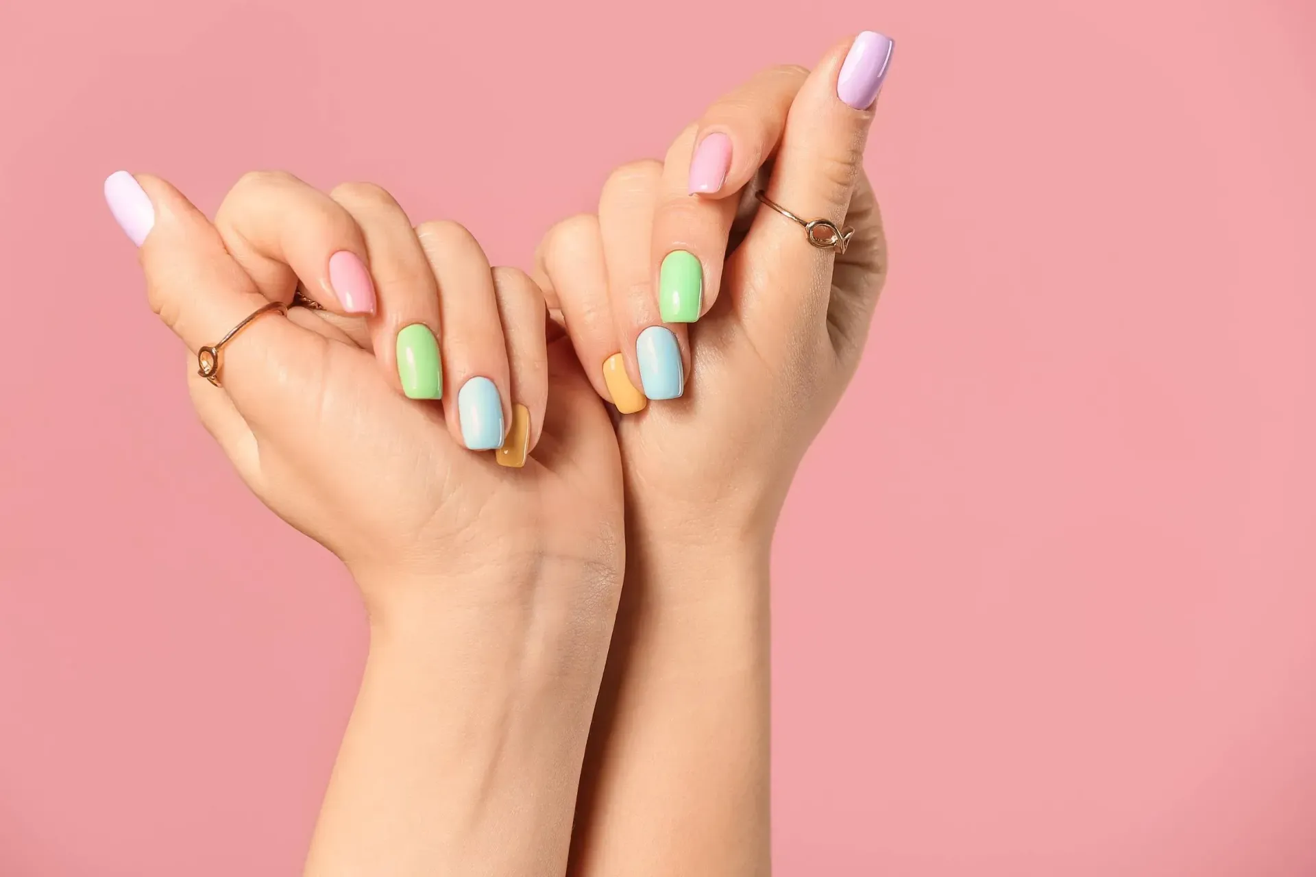 Manos de mujer con uñas de colores y anillos sobre un fondo rosa.
