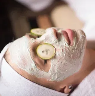 Una mujer se aplica una mascarilla de arcilla en la cara con rodajas de pepino en los ojos.