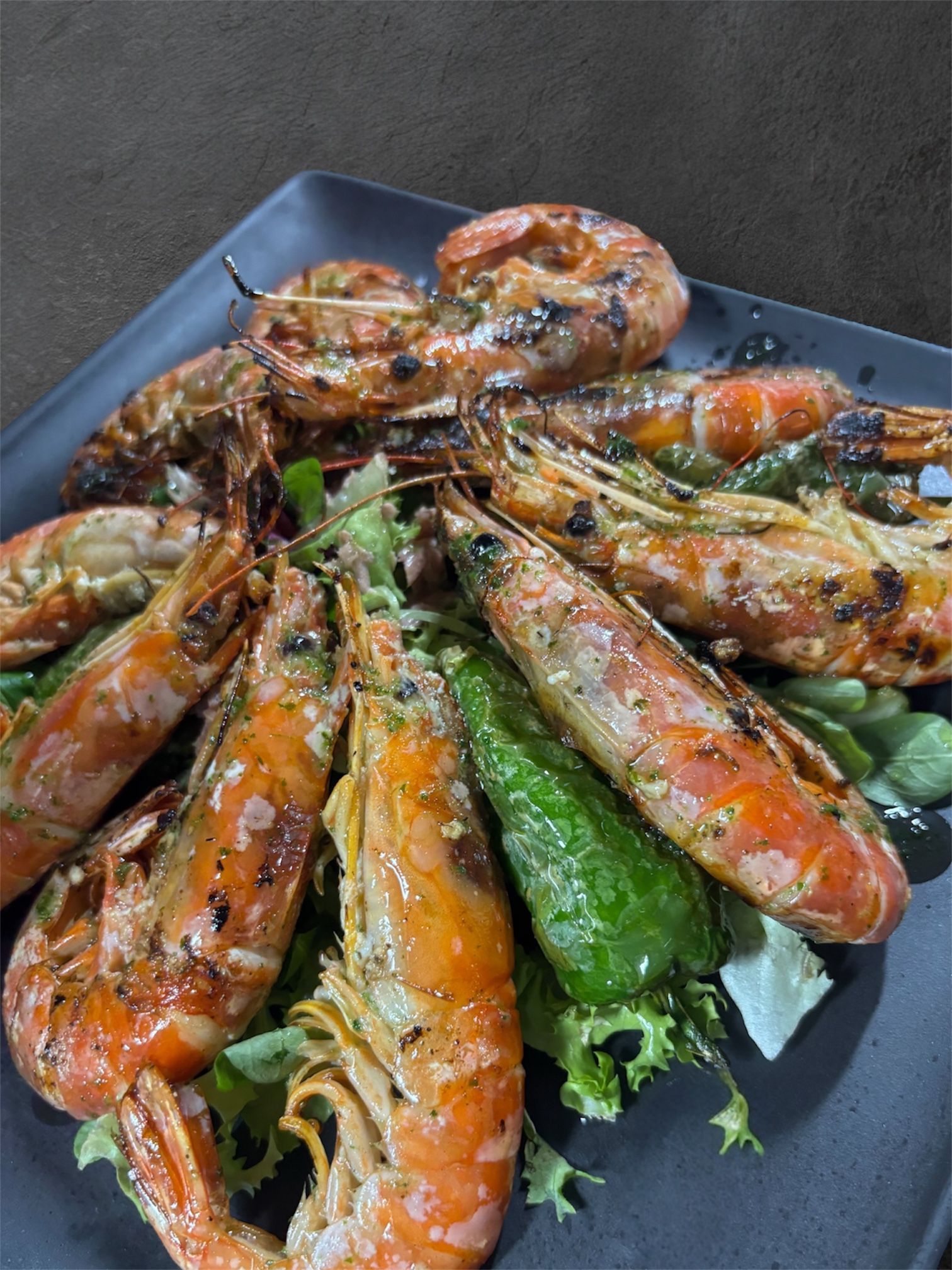 Camarones a la plancha sobre una cama de lechuga, con pimiento verde, en un plato cuadrado negro.