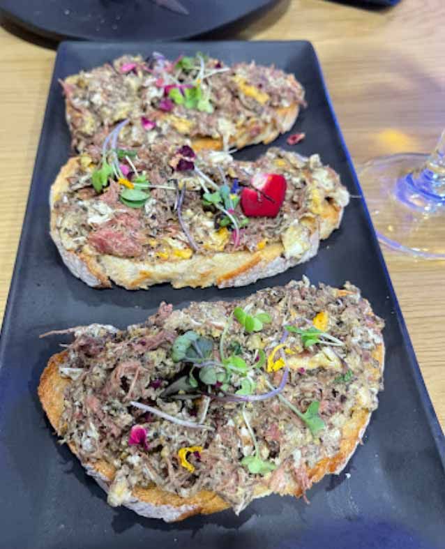 Tres piezas de comida en un plato negro con una copa de vino al fondo