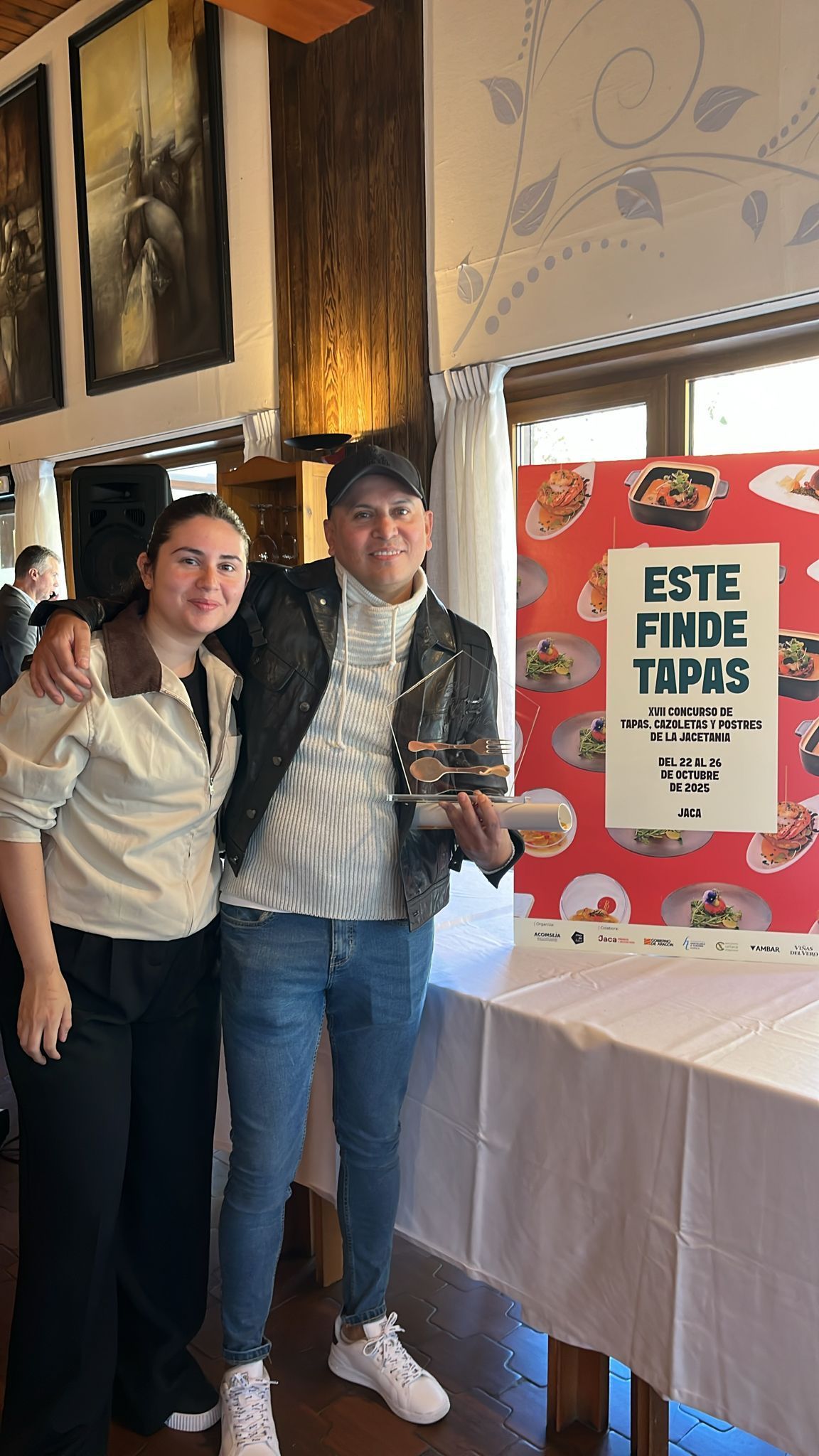Dos personas están junto a un anuncio de tapas. La persona de la derecha sostiene un premio. Sonríen en un restaurante.