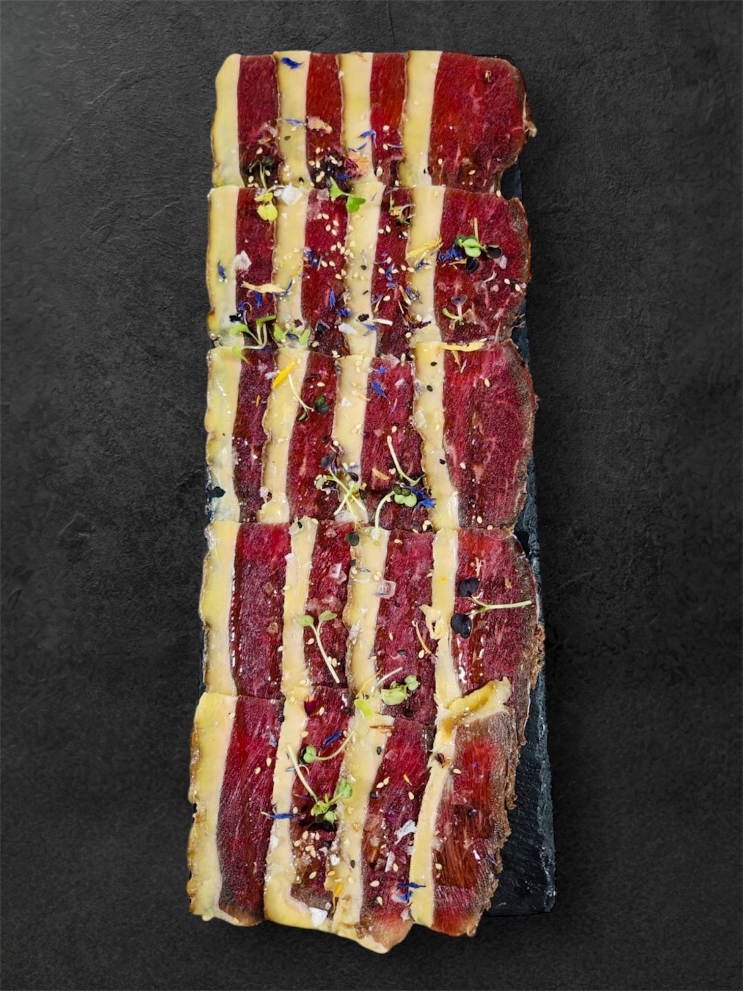 Aperitivo de carne cortada en rodajas sobre una superficie oscura, alternando rayas rojas y pálidas, adornado con hierbas.