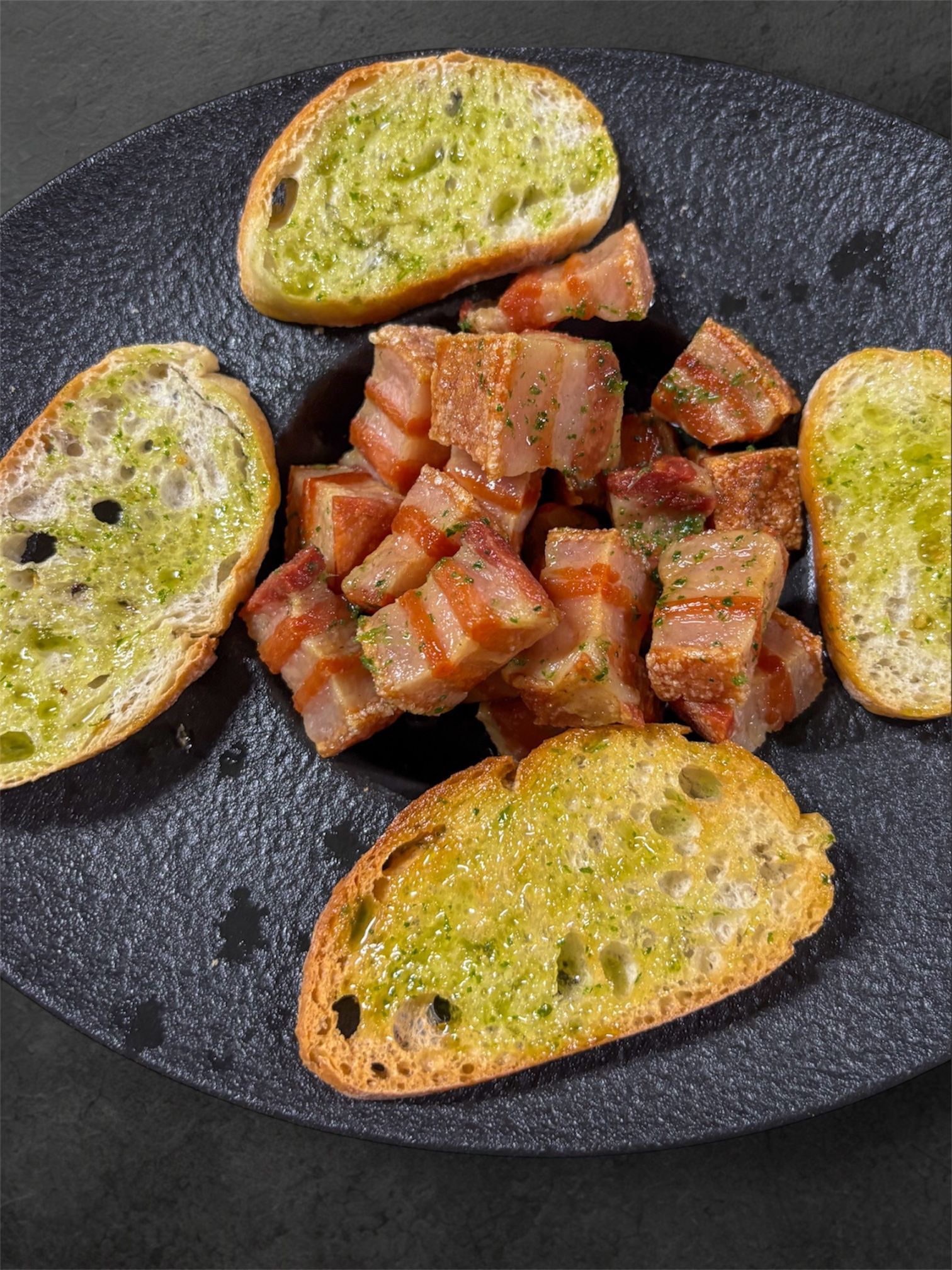 Cubitos de tocino y rebanadas de pan tostado con cobertura verde en un plato oscuro.