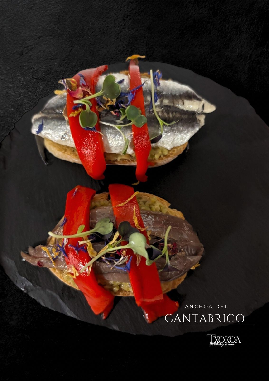 Dos tapas sobre pizarra: anchoas, pimientos rojos y microgreens sobre pan tostado.