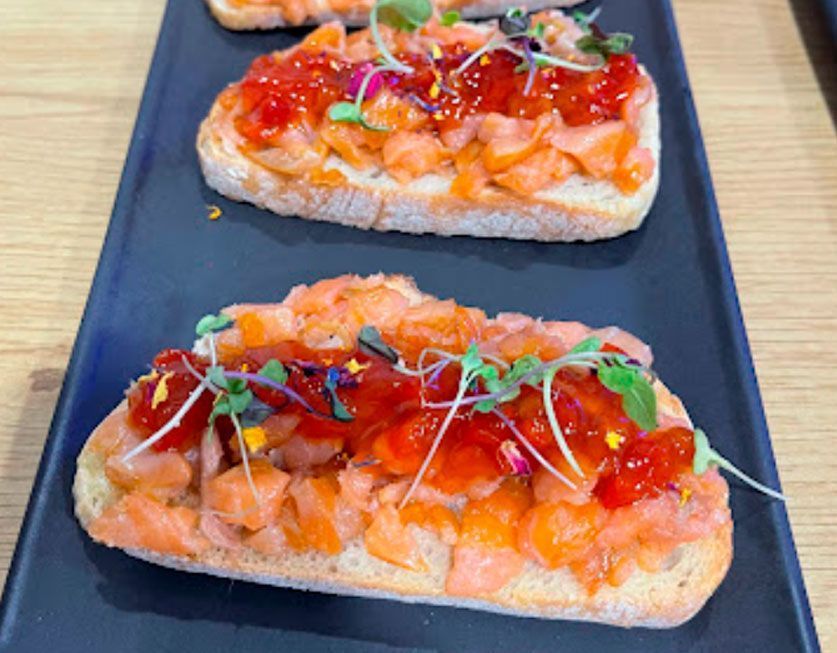Tres sándwiches con salmón y tomates en un plato negro sobre una mesa.