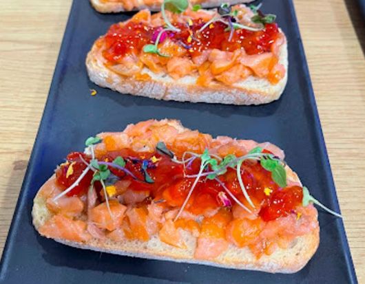 Tres sándwiches con salmón y tomates en un plato negro sobre una mesa.