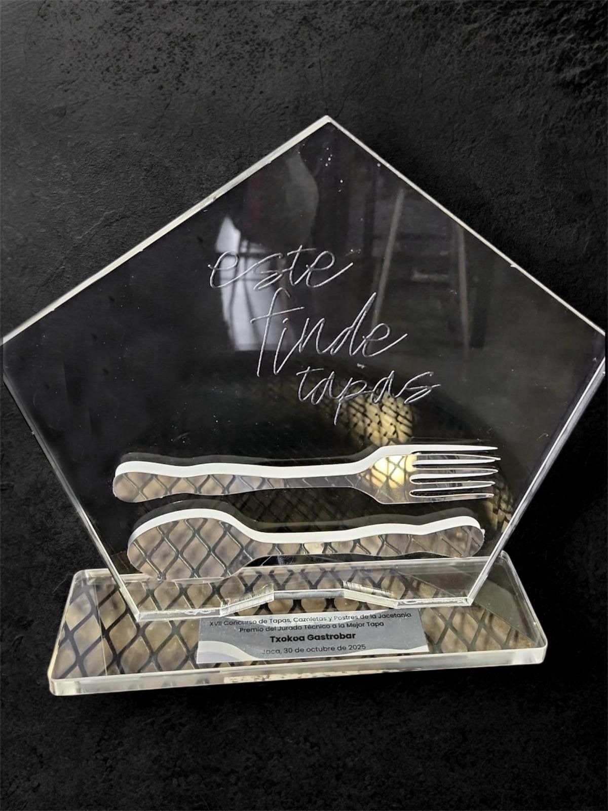 Trofeo de acrílico transparente con texto grabado, tenedor y cuchara, y una placa de identificación sobre un fondo oscuro.