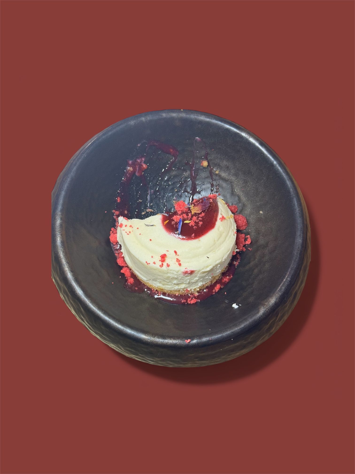 Rebanada de tarta de queso en un recipiente oscuro, salsa roja, migas sobre fondo rojo.