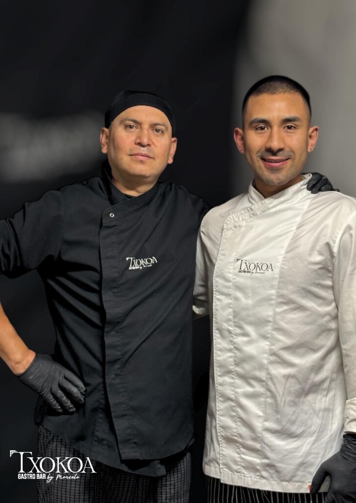 Dos chefs con batas blancas y negras, de pie frente a un fondo oscuro. Sonríen.