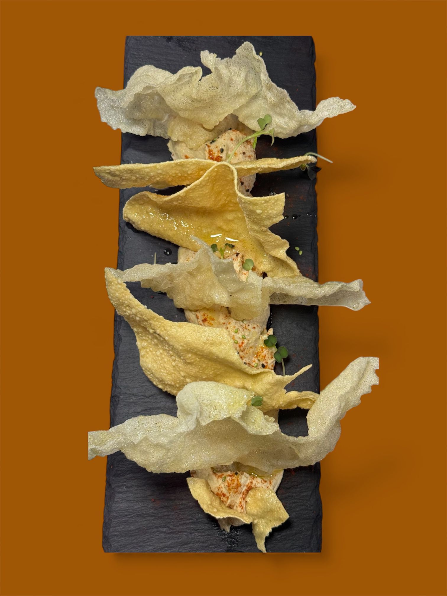 Aperitivo deconstruido con patatas fritas doradas e infladas dispuestas sobre una salsa cremosa en una bandeja rectangular negra.