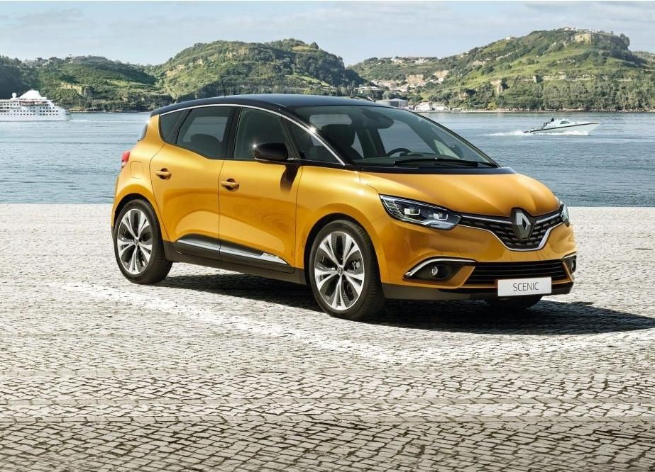 Une Renault orange est garée sur un quai pavé avec des collines et des bateaux en arrière-plan.