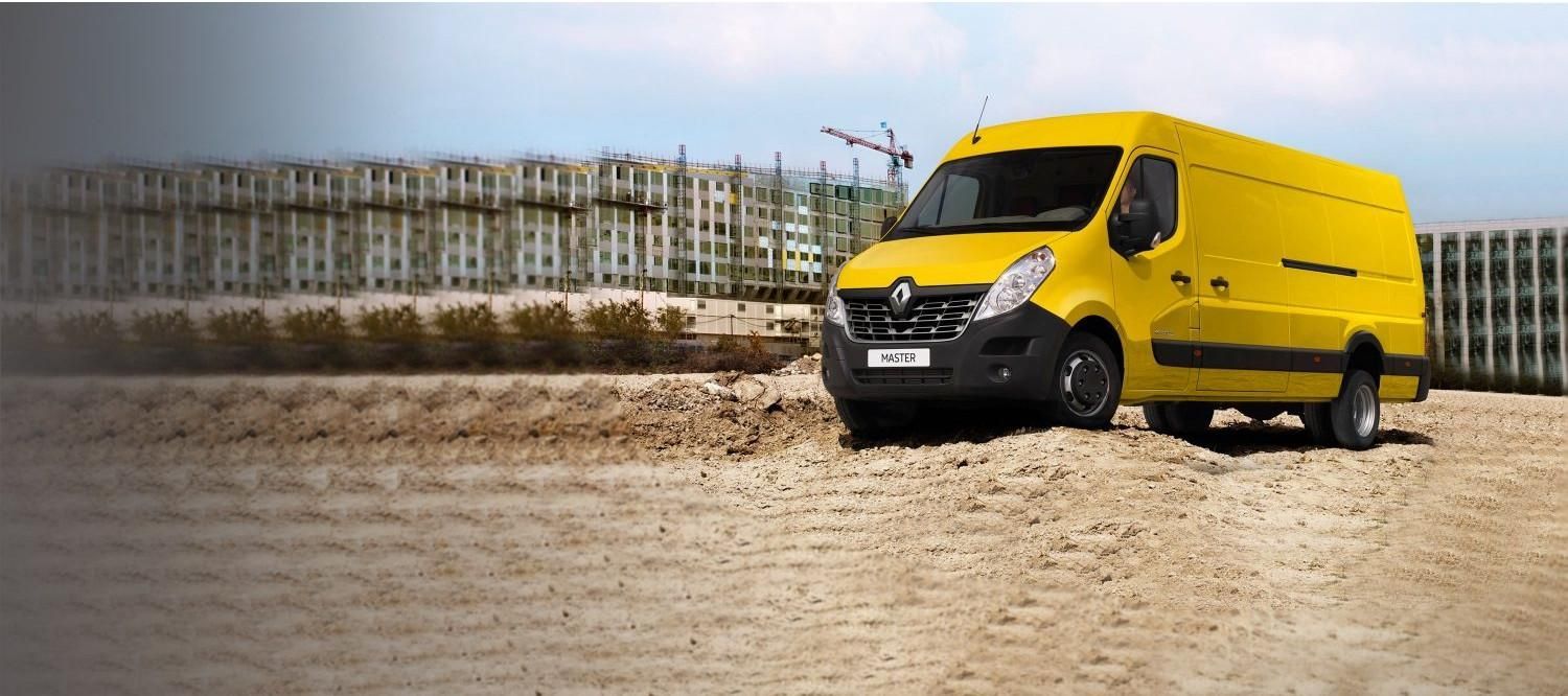 Une camionnette jaune circule sur un terrain sablonneux avec des immeubles en arrière-plan.