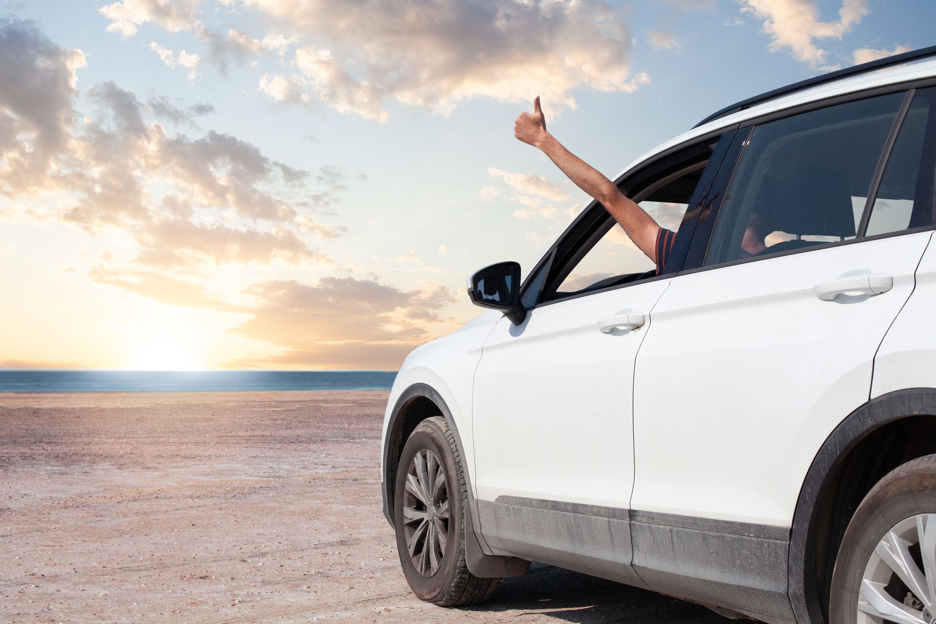 Un SUV blanc sur une plage au coucher du soleil. Un homme passe son bras par la fenêtre et lève le pouce en l'air.