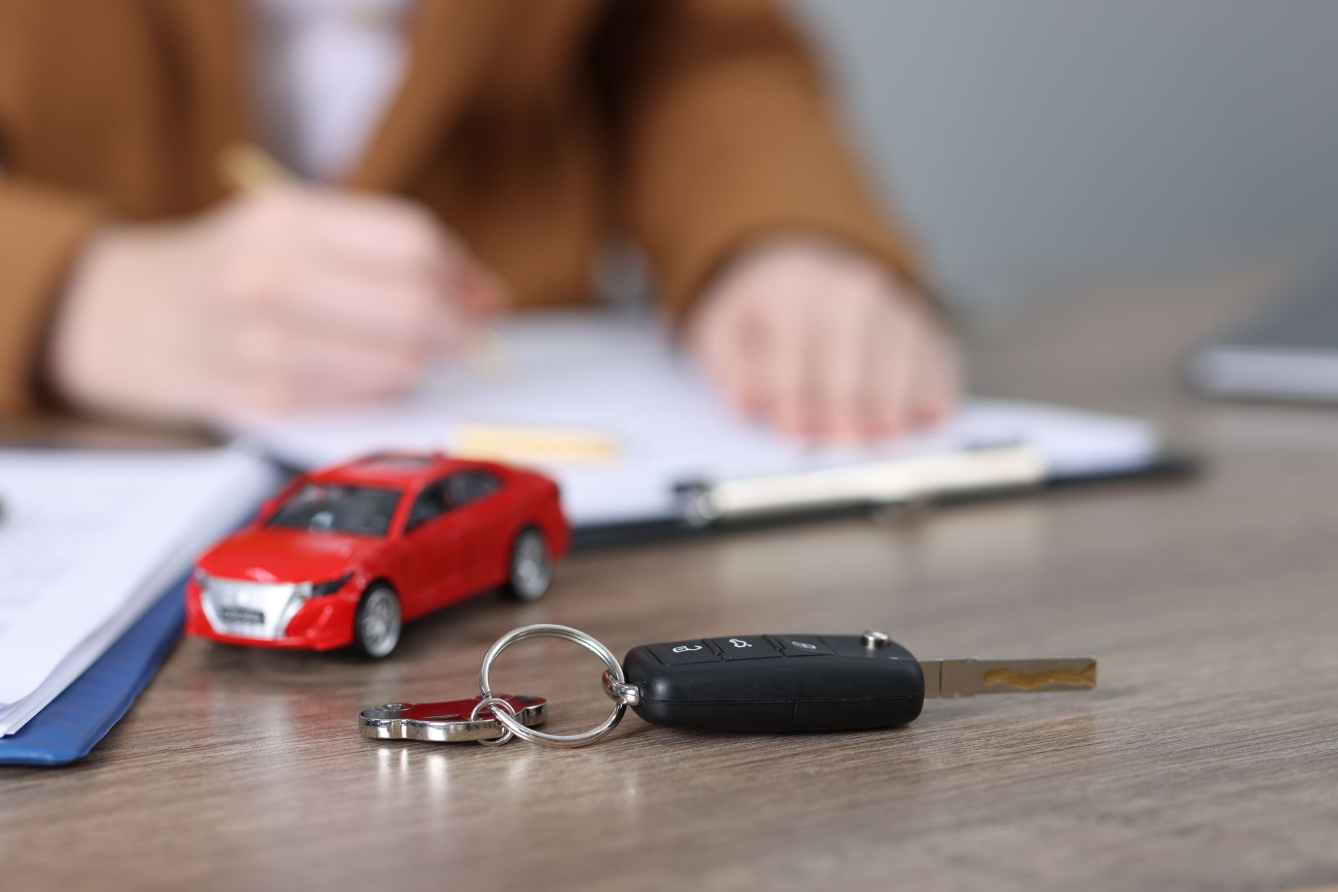 Une voiture miniature et des clés sur un bureau, avec une personne floue écrivant en arrière-plan.