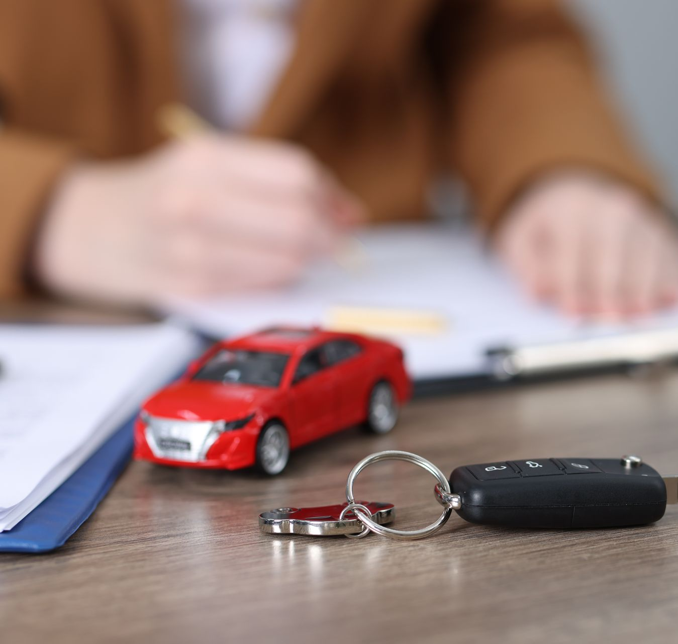 Une petite voiture rouge et un porte-clés noir sur un bureau, avec une personne floue écrivant en arrière-plan.
