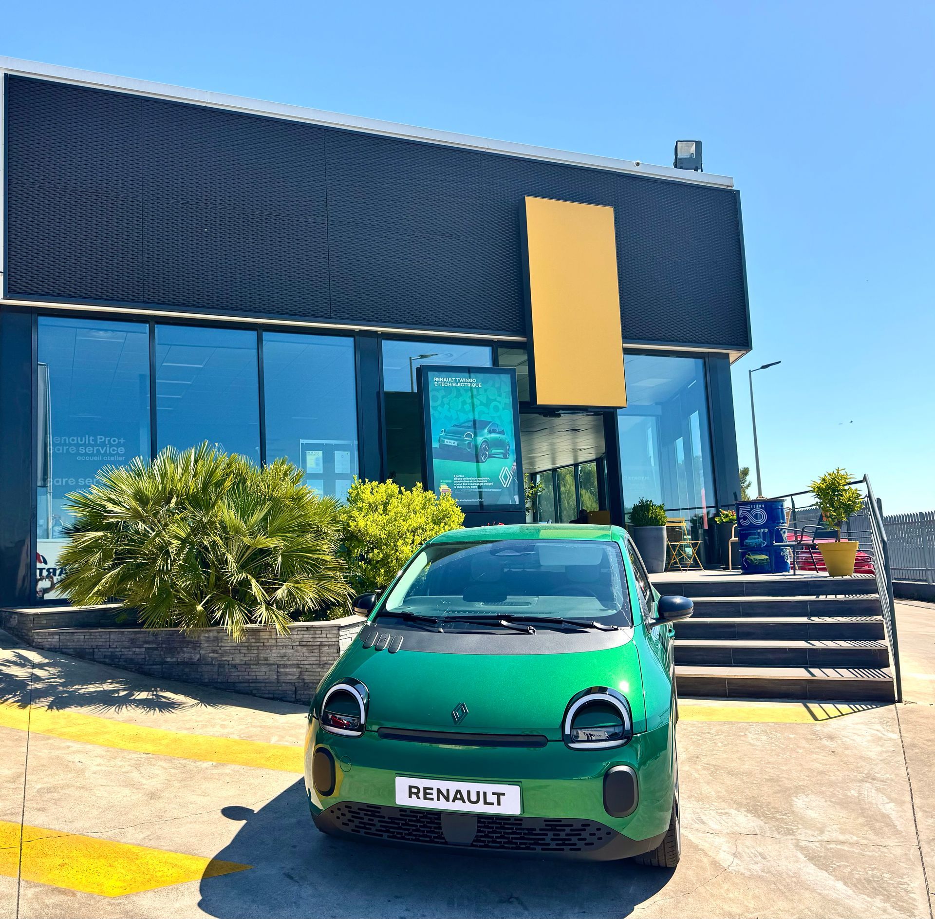 Une Renault Twingo est garée devant une concession aux baies vitrées ensoleillées.