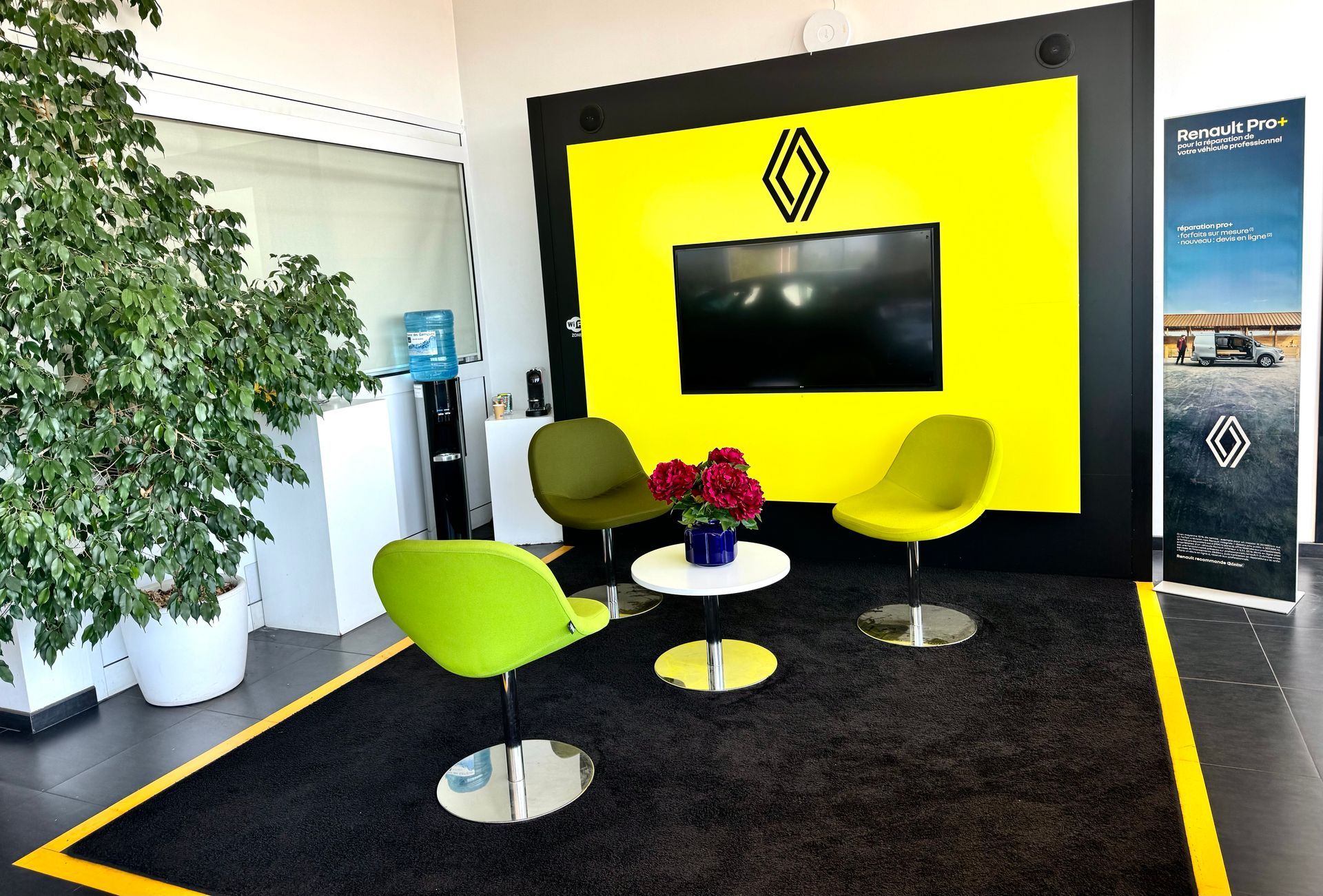 Salle d'attente jaune dans une concession Renault, avec un écran de télévision mural, des chaises et une table basse.