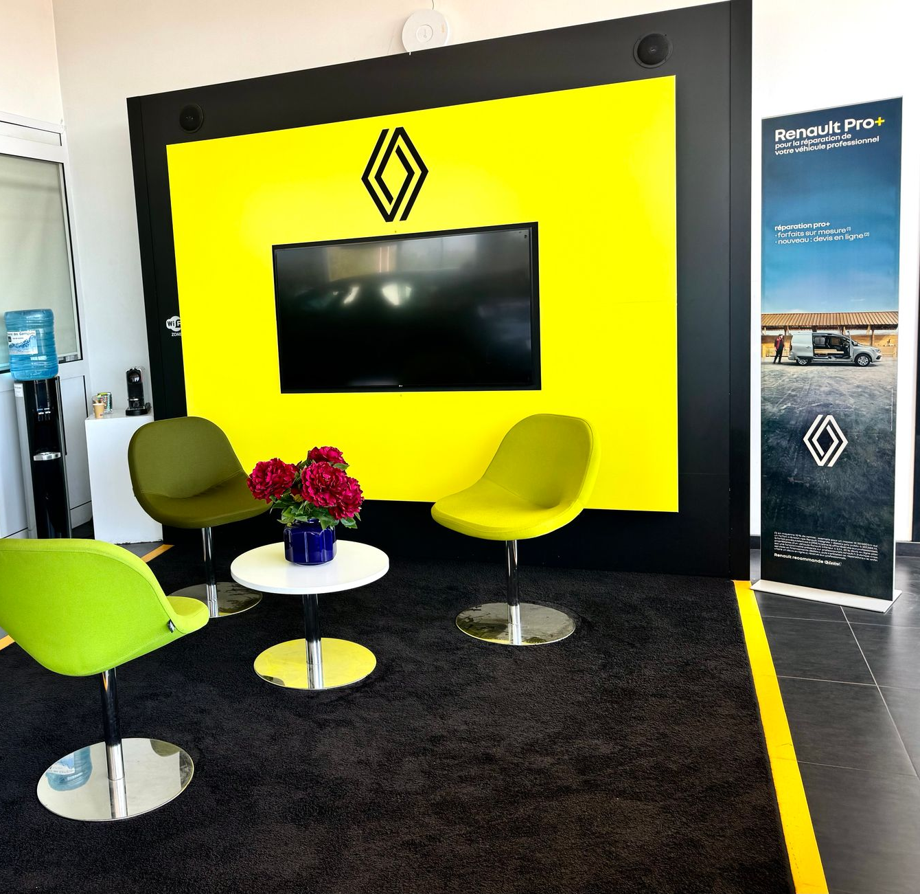 Salon d'accueil moderne avec mur jaune vif orné du logo Renault, télévision murale, chaises et bouquet de fleurs sur une table basse.