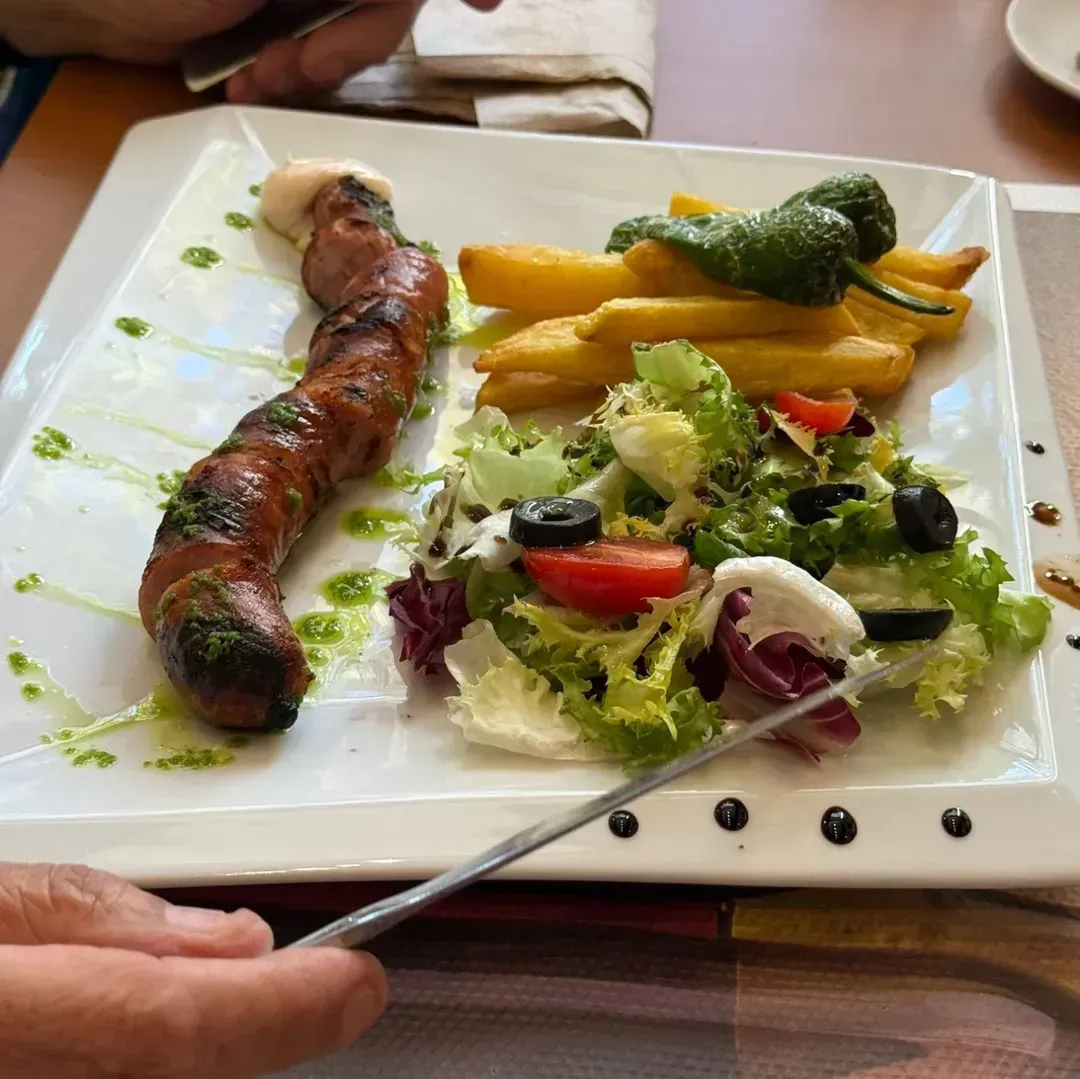 Una salchicha a la parrilla servida con papas fritas, un pimiento verde y una ensalada en un plato blanco