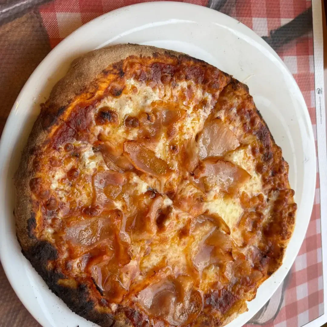 Una pizza redonda, cocinada en horno de leña, con queso fundido y jamón, servida en el centro de un plato blanco
